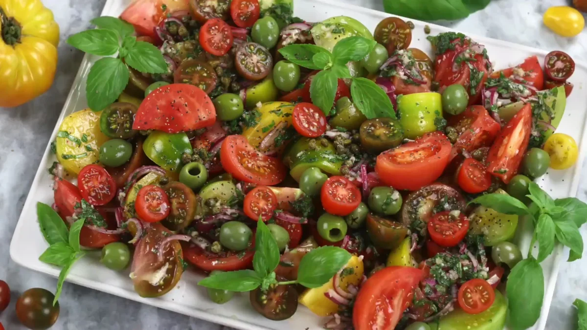 Heirloom Tomato Salad 10 Serveing A fresh tomato salad with sliced tomatoes, green herbs, and a sprinkle of seasoning, presented in a wooden bowl.